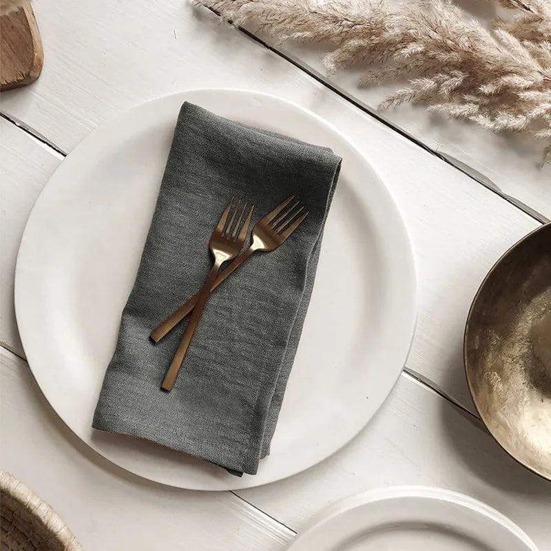 Une assiette blanche avec une serviette grise et des couverts en métal doré posés sur une table en bois, avec des éléments décoratifs en arrière-plan.
