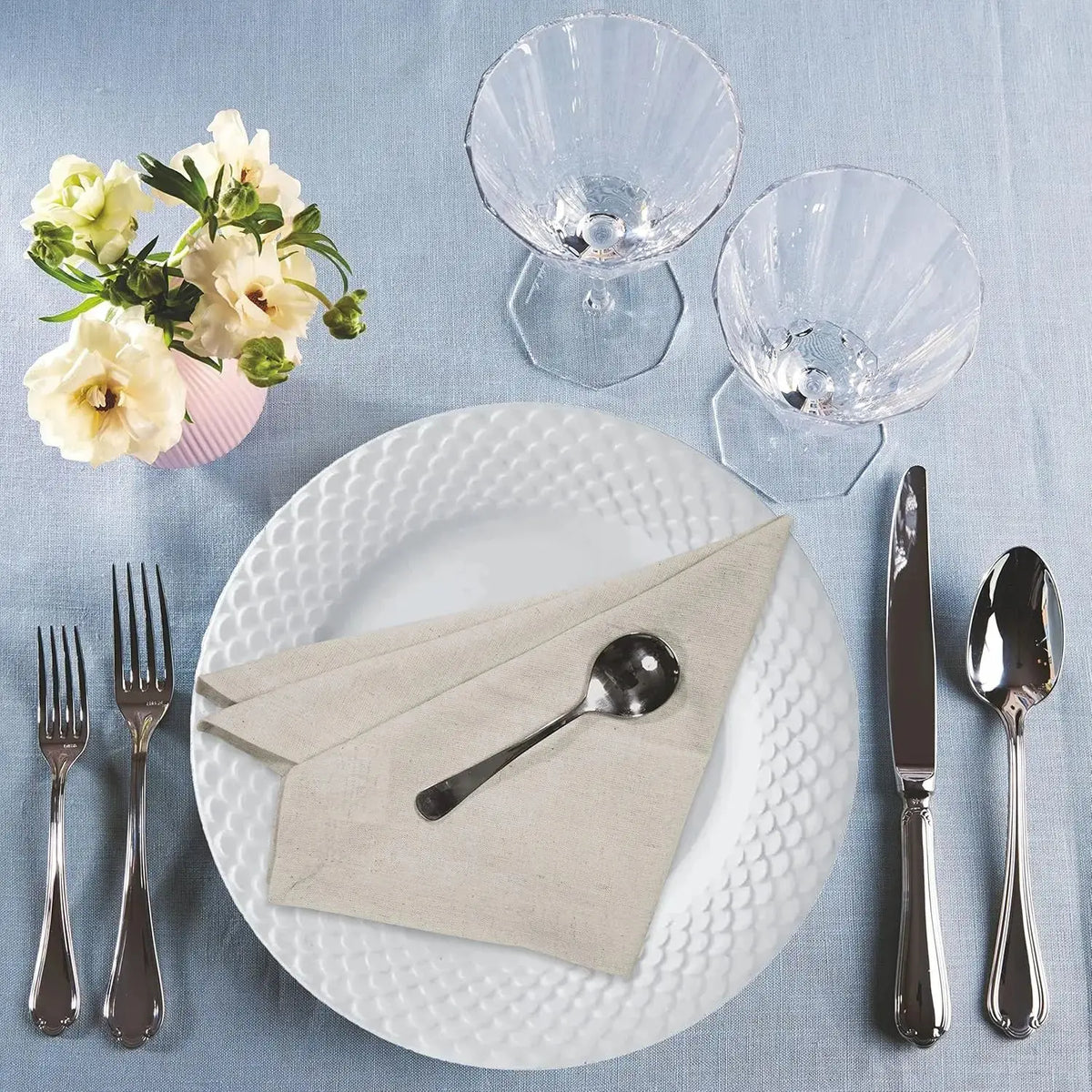 Cette image montre une élégante configuration de table de restaurant avec des assiettes et des verres en cristal, des couverts en métal brillant et un bouquet de fleurs blanches dans un vase rose sur fond gris.