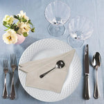 Cette image montre une élégante configuration de table de restaurant avec des assiettes et des verres en cristal, des couverts en métal brillant et un bouquet de fleurs blanches dans un vase rose sur fond gris.
