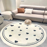 Tapis rond en peluche beige décoré de motifs de coeurs noirs, placé sur un sol dans un salon avec un canapé gris, des coussins et une peluche sur une table basse.