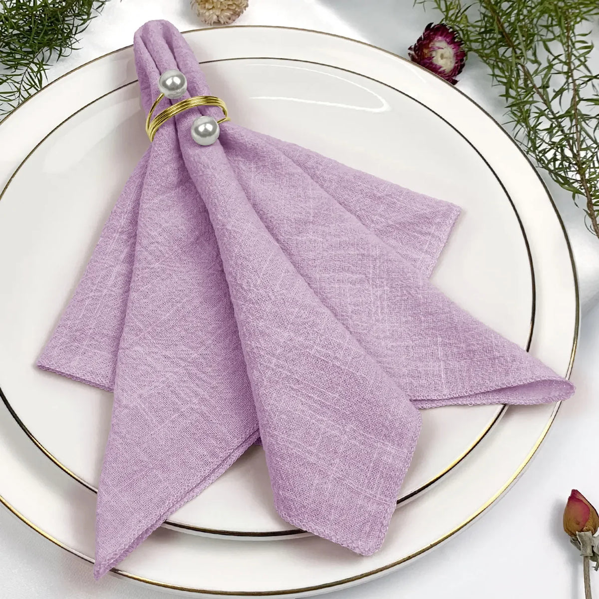 Une serviette de table violette ornée de perles dorées repose sur une assiette blanche entourée de fleurs et de feuillages.