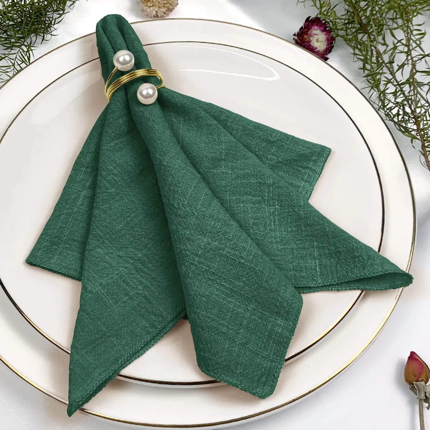 Un magnifique pli de serviette en tissu vert foncé, orné de perles blanches et entouré de fleurs décoratives sur un plateau blanc.
