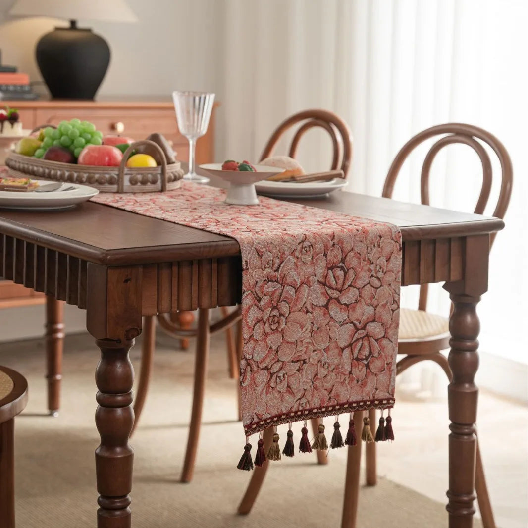Une table à manger en bois avec un chemin de table fleuri et décoratif, des assiettes et des fruits disposés dessus, dans un intérieur lumineux.