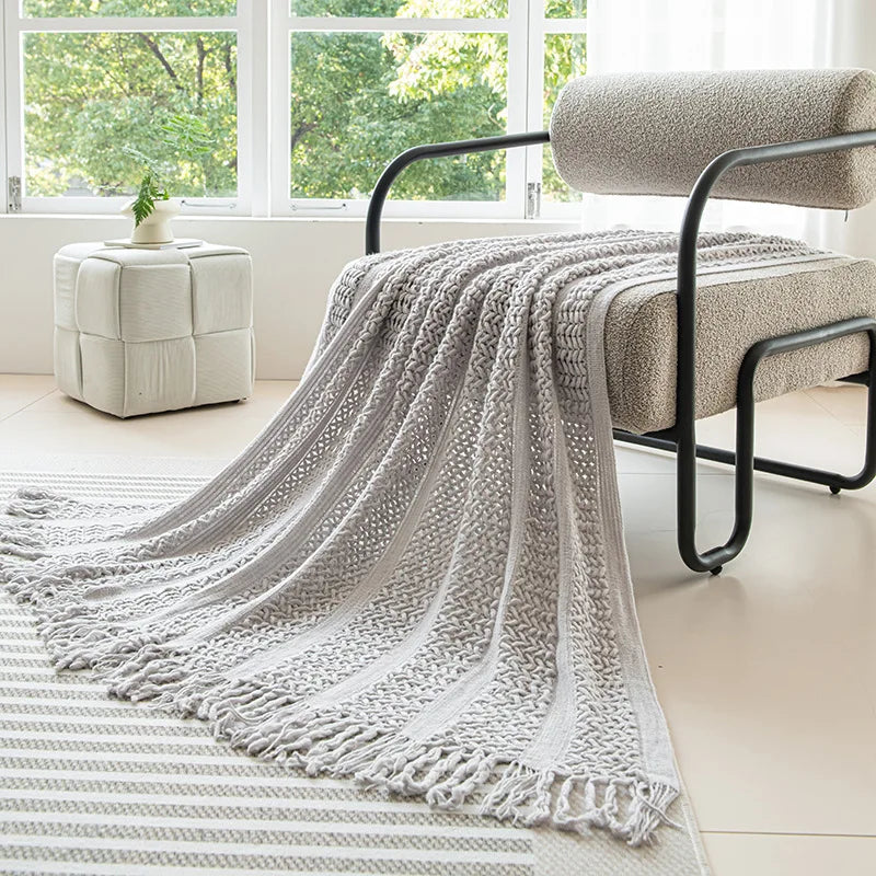 Une couverture en tricot grise élégante et confortable, posée sur un fauteuil dans un salon lumineux avec une vue sur les arbres à l'extérieur.