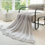 Une couverture en tricot grise élégante et confortable, posée sur un fauteuil dans un salon lumineux avec une vue sur les arbres à l'extérieur.