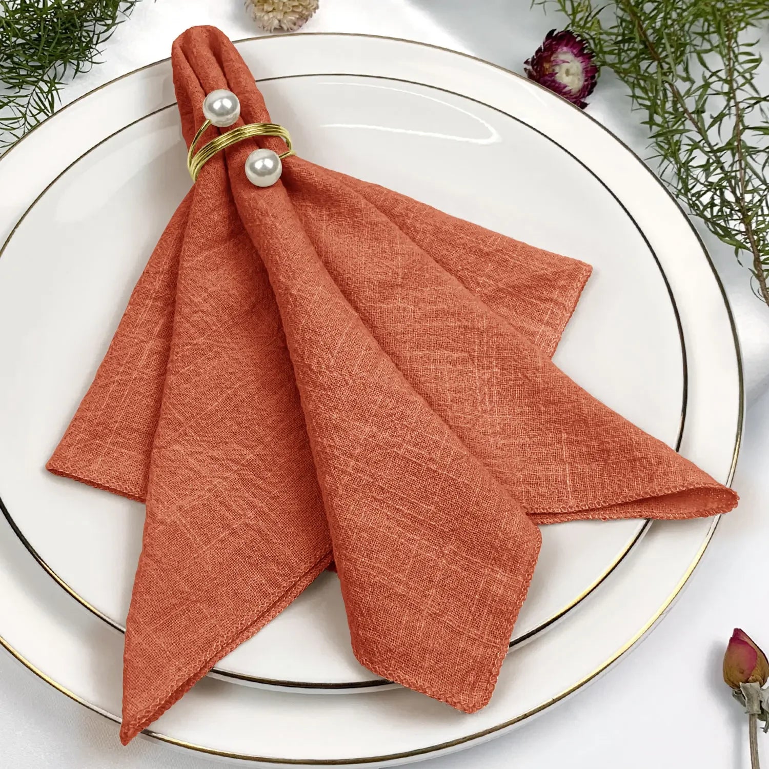 Une serviette de table pliée de couleur orange, ornée de perles blanches, posée sur une assiette blanche entourée de décorations végétales.