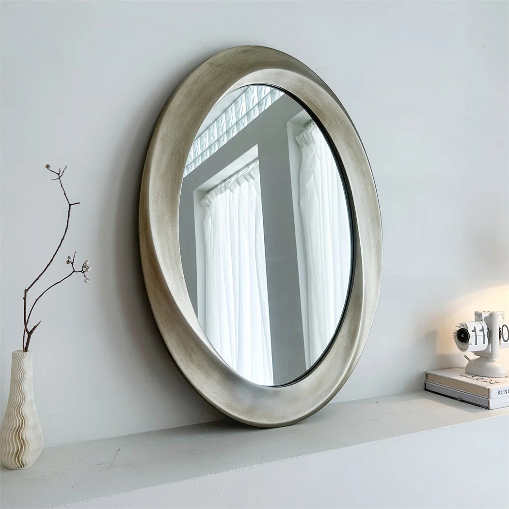 Le miroir ovale gris clair reflète élégamment les rideaux blancs et les éléments décoratifs sur l'étagère, créant une atmosphère apaisante et harmonieuse dans la pièce.