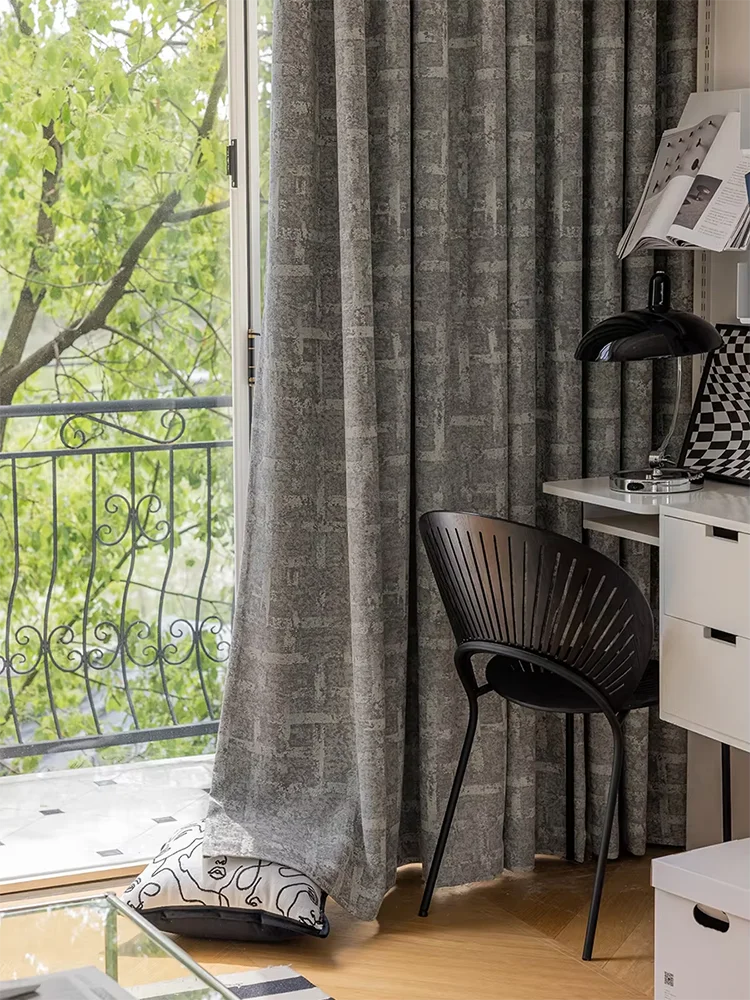 Rideaux gris élégants dans un intérieur moderne, apportant une ambiance raffinée à une pièce lumineuse avec vue sur un balcon verdoyant. Belle chaise noire design et bureau minimaliste, parfaits pour un espace de travail créatif.