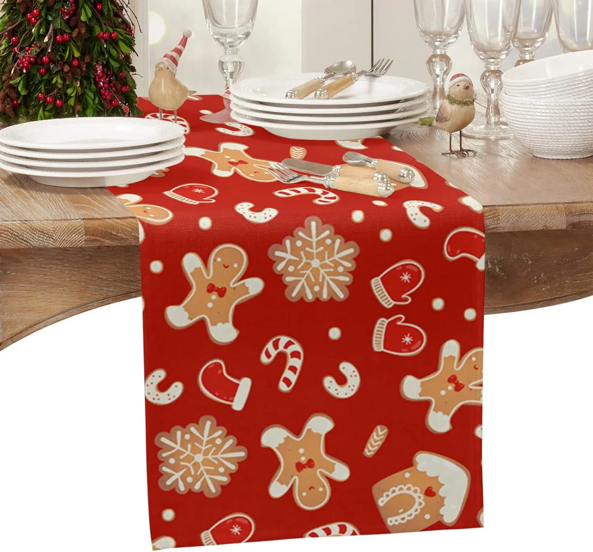 Une table de fête décorée pour Noël avec de la vaisselle blanche, des verres de cristal et un chemin de table rouge orné de motifs de bonhommes en pain d'épice et de flocons de neige.