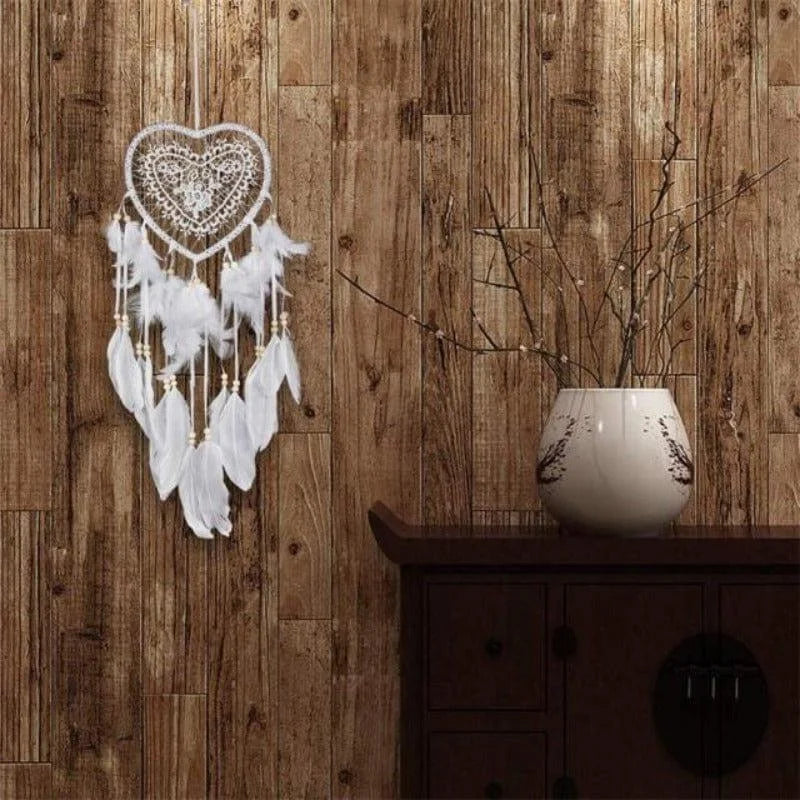 Attrape-rêves en forme de cœur avec plumes blanches suspendu sur un mur en bois rustique, à côté d'un vase décoratif moderne sur une commode. Parfait pour une décoration intérieure apaisante et élégante, ajoutant une touche bohème à votre espace de vie.