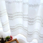 Rideau décoratif blanc avec motifs floraux délicats et bordure festonnée. Parfait pour ajouter une touche élégante et lumineuse à votre intérieur. Accompagne un cadre photo et un bouquet sur une surface brillante. Idéal pour un salon ou une chambre.