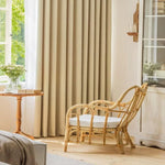 Fauteuil en rotin au design élégant dans un salon lumineux avec des rideaux beiges, une table d'appoint avec des livres et un vase de fleurs, créant une atmosphère chaleureuse et accueillante.