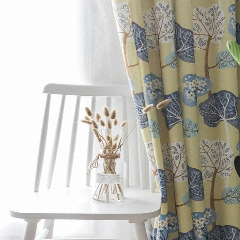 Rideau décoratif avec motif de feuilles bleues et beiges accroché devant une chaise blanche, dans une ambiance lumineuse et moderne. Accessoirisé avec un vase en verre et un bouquet naturel pour un style élégant et apaisant, idéal pour embellir votre intérieur.