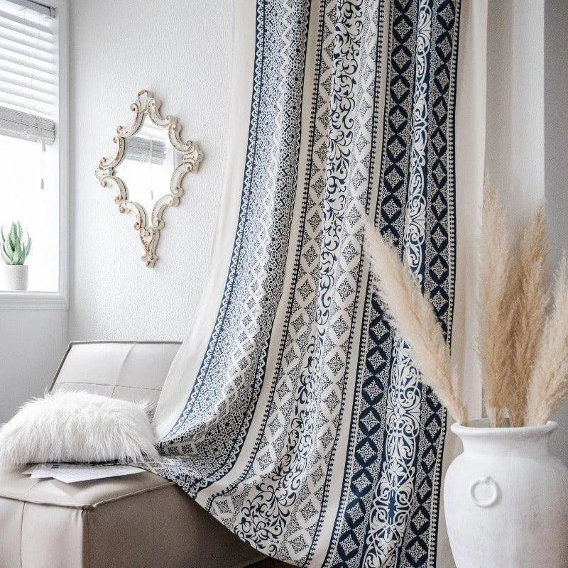 Rideau élégant avec motifs graphiques bleu et blanc suspendu dans un salon lumineux. Un design chic qui ajoute une touche de sophistication à tout intérieur moderne. Accessoires décoratifs visibles : coussin duveteux, pot de plante, herbes séchées, et miroir mural ornemental. Parfait pour créer une ambiance cosy et accueillante.