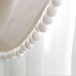 Voilage décoratif blanc avec détails de pompons, idéal pour embellir les fenêtres de votre intérieur et apporter une touche d'élégance douce et moderne.