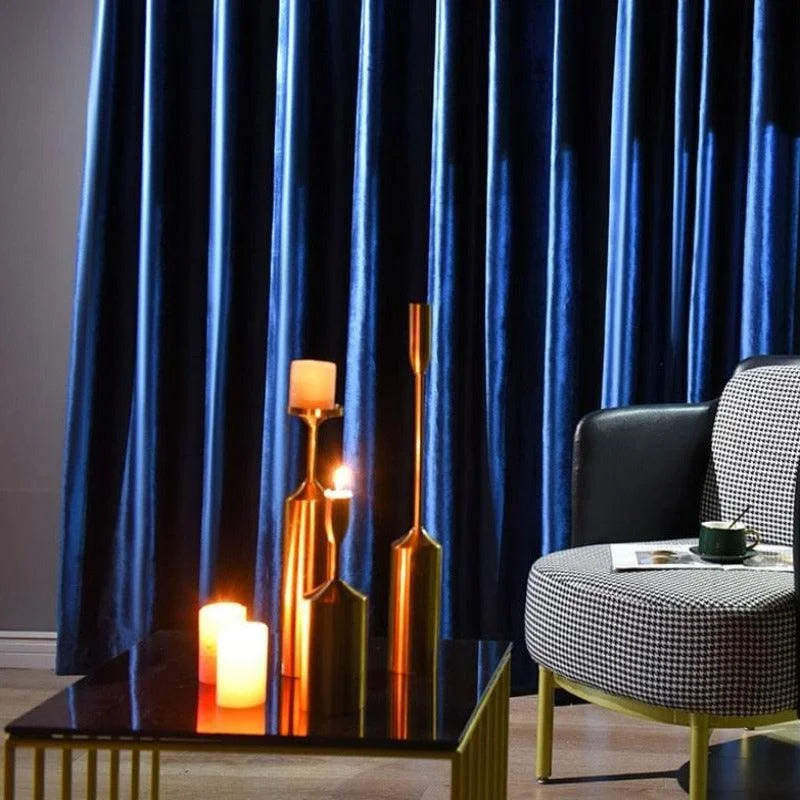 Rideaux élégants bleus foncés dans un salon moderne, accentués par des chandeliers dorés et des bougies sur une table basse. Ambiance chaleureuse et contemporaine avec un fauteuil confortable et une tasse de café. Idéal pour apporter une touche de luxe et de confort à votre intérieur.