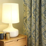 Lampe de table moderne avec abat-jour blanc posée sur une table en bois clair, accompagnée d'un rideau à motifs élégants en arrière-plan. Idéale pour ajouter une touche de sophistication à votre salon ou chambre à coucher. Accessoires de décoration discrets mais chic visibles sur la table.