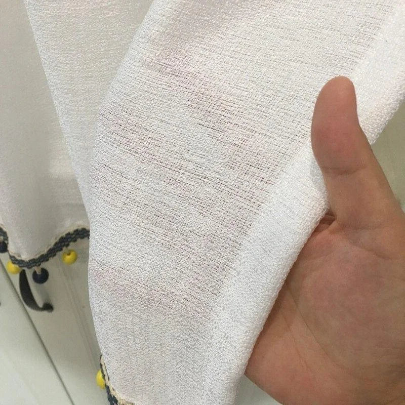 Voile blanc semi-transparent avec bordure décorative en pompons jaunes et bleus, idéal pour ajouter une touche élégante à votre décoration intérieure. Parfait pour les rideaux ou autres projets créatifs dans votre maison.