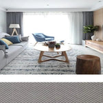 Salon moderne et lumineux avec canapé gris, fauteuil bleu et table basse en bois, accentués par un tapis au motif géométrique. Ambiance chaleureuse grâce aux accessoires décoratifs et à la luminosité naturelle provenant de la grande baie vitrée. Idéal pour une décoration intérieure contemporaine et épurée.