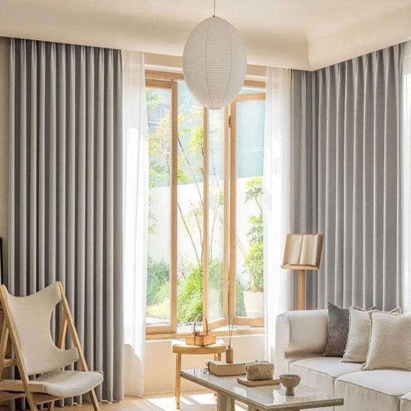 Salon moderne et lumineux avec de grands rideaux élégants gris, offrant une ambiance chaleureuse et douce. Une belle lumière naturelle inonde la pièce grâce aux grandes fenêtres. Mobilier minimaliste et contemporain, avec un canapé blanc confortable, une chaise design et une table basse épurée. Accessoires décoratifs soigneusement disposés pour une atmosphère accueillante et harmonieuse.