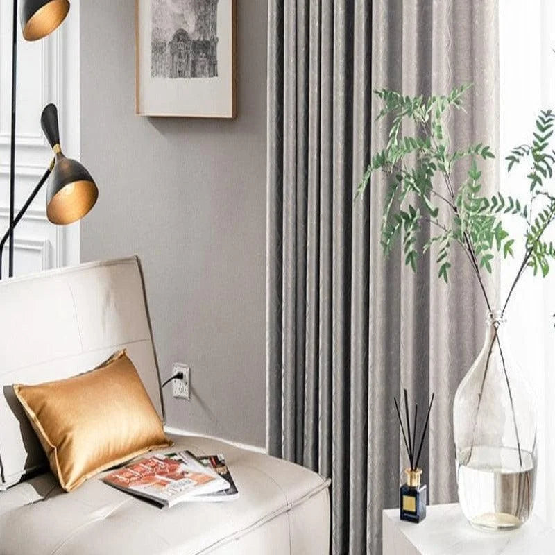 Un coin de salon élégant avec un canapé blanc moderne rehaussé par un coussin doré, éclairé par une lampe design à trois ampoules noires et dorées. Les rideaux gris ajoutent une touche de sophistication tandis qu'un vase transparent avec des feuilles vertes et un diffuseur sur une table blanche complètent la décoration intérieure minimaliste. Idéal pour un intérieur chic et raffiné.
