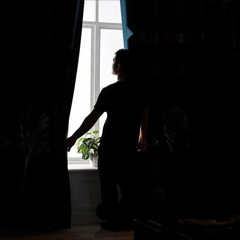 Homme debout près d'une fenêtre, ouvrant un rideau pour laisser entrer la lumière dans une pièce sombre, avec une plante en pot sur le rebord de la fenêtre, créant une ambiance apaisante et naturelle.