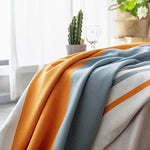 Couverture décorative aux couleurs vives orange, bleu et blanc, posée sur un meuble dans un intérieur lumineux avec une plante verte et un cactus en arrière-plan, idéale pour ajouter une touche moderne et chaleureuse à votre salon.