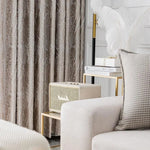 Image d'un salon élégant avec rideaux texturés marron, un canapé confortable et un coussin à motifs. Une lampe stylisée et des livres sur une table d'appoint dorée ajoutent une touche de sophistication. Une enceinte vintage complète l'ambiance chic et moderne.