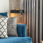 Canapé bleu élégant avec coussins décoratifs dans un salon moderne. Éclairage doux d'une lampe sophistiquée et rideaux gris pour une ambiance chaleureuse. Design intérieur contemporain mettant en valeur le confort et le style.