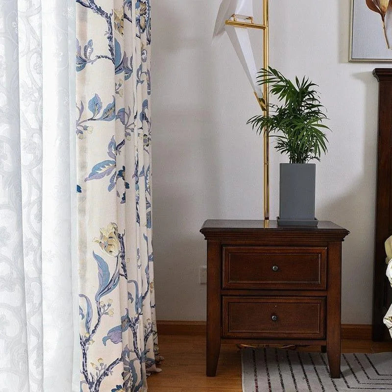 Chevet en bois marron avec tiroirs dans une chambre élégante, agrémentée de rideaux floraux bleus et blanc cassé, une décoration moderne avec une plante verte et un lampadaire doré, ajoutant une touche de raffinement à l'espace. Idéal pour les intérieurs sophistiqués et chaleureux.