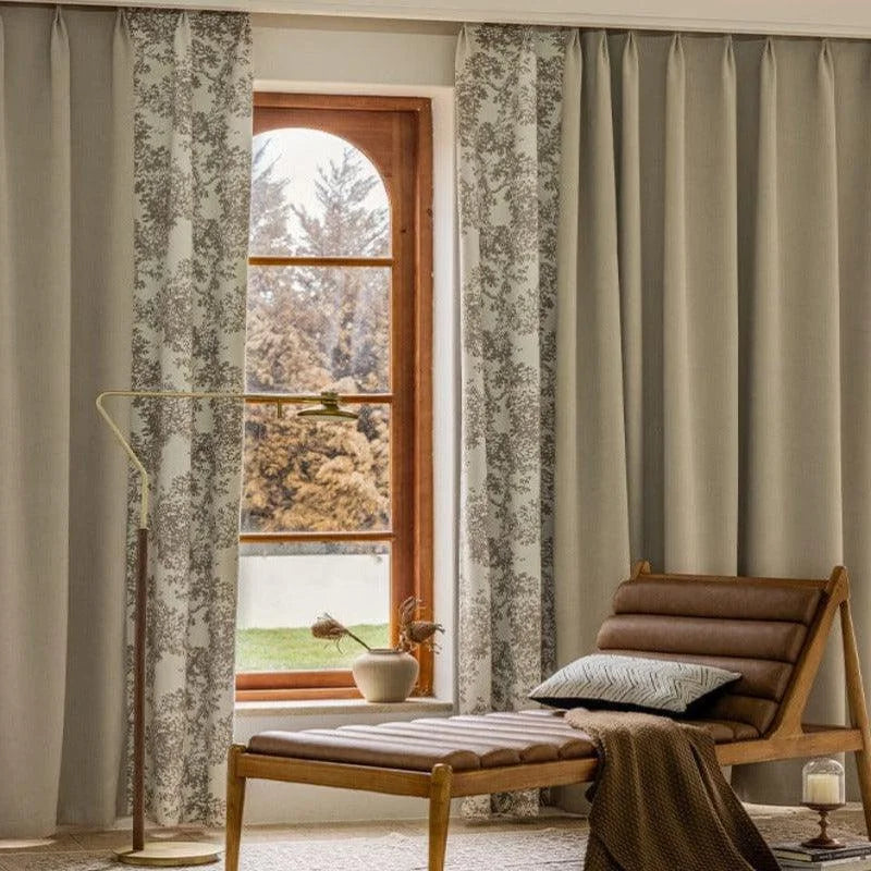 Chaise longue design dans un salon élégant avec grandes baies vitrées. Rideaux élégants avec motifs floraux offrant une ambiance chaleureuse. Luminaire moderne en métal doré à côté d'un vase décoratif. Espace intérieur lumineux et confortable avec vue extérieure sur jardin.