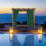 Pergola avec rideaux vert foncé sur terrasse élégante avec vue sur mer au coucher du soleil. Éclairage romantique et ambiance détente autour de la piscine, idéale pour les soirées extérieures. Aménagement extérieur sophistiqué pour jardin ou terrasse, alliant confort et esthétique.