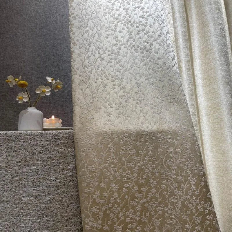 Rideau élégant crème avec motif floral en relief, idéal pour salon ou chambre, apportant une touche de sophistication et de douceur à votre intérieur. Accessoirisé d'un vase blanc contenant des fleurs et une bougie allumée pour compléter l'ambiance.