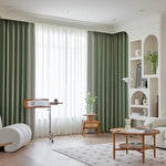 Salon moderne avec rideaux verts et décor minimaliste. Espace lumineux grâce à une grande fenêtre. Mobilier élégant comprenant une table basse en bois, étagères intégrées et fauteuil confortable. Parfait pour une ambiance chaleureuse et accueillante. Design intérieur contemporain avec touches de déco naturelle.