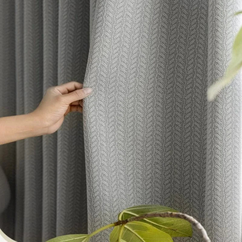 Rideau gris élégant avec motif géométrique, idéal pour une décoration intérieure moderne. Ce rideau offre une ambiance apaisante et chic à votre salon ou chambre. Personne ajustant le rideau, ajoutant une touche de sophistication à votre espace de vie. Plante verte en arrière-plan, soulignant un style naturel et harmonieux.