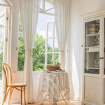 Table ronde en bois élégante et accessoires en osier, parfaits pour une décoration intérieure lumineuse. Profitez de l'ambiance chaleureuse grâce à des rideaux longs blancs laissant passer la lumière naturelle. Ideal pour un salon chic et aéré.