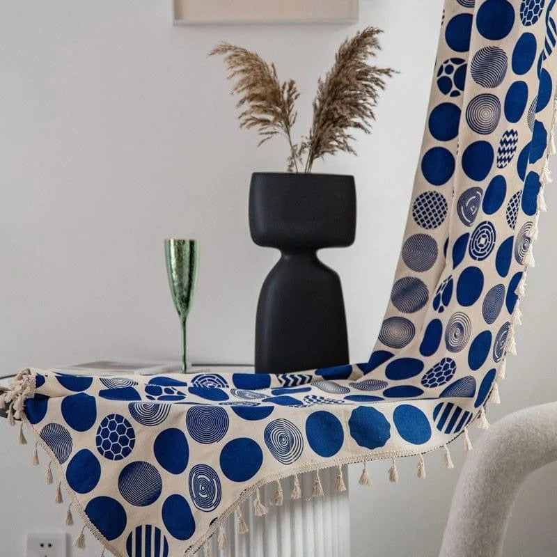 Tapisserie décorative avec motifs géométriques bleus et franges, posée sur une table claire. Décoration intérieure élégante avec vase noir moderne et verre décoratif vert, idéale pour un salon contemporain. Accessoire de maison tendance ajoutant une touche artistique et vibrante à votre décoration intérieure.