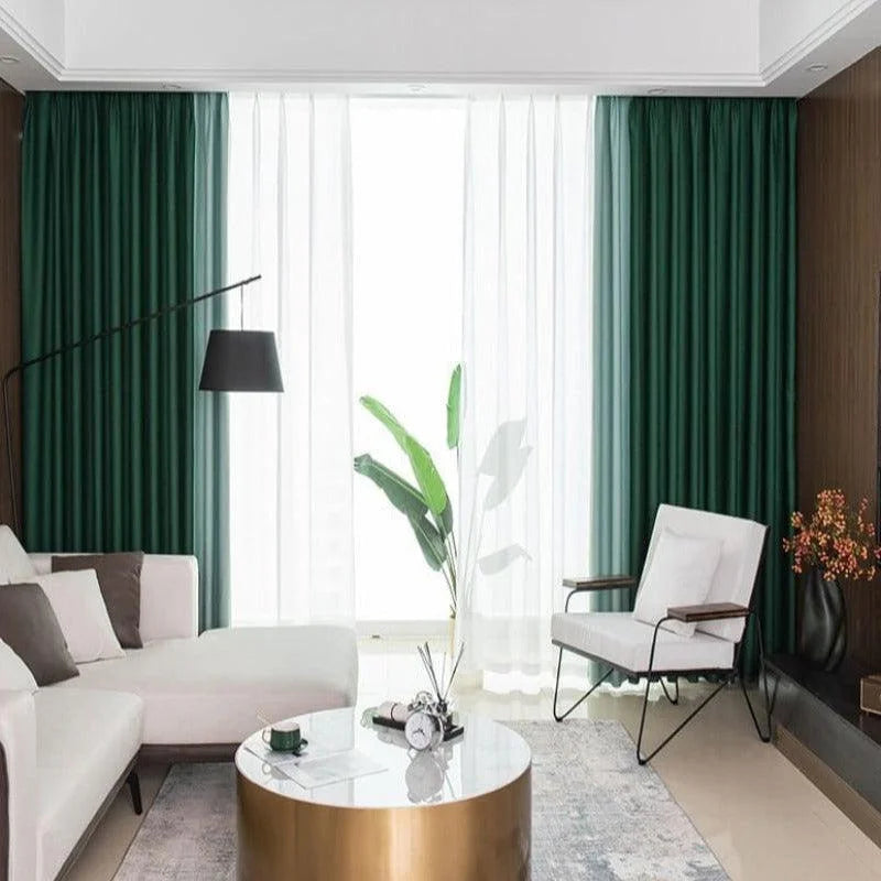 Salon moderne avec canapé d'angle blanc et fauteuil élégant. Rideaux verts sombres et transparents encadrant une grande fenêtre pour un espace lumineux. Table basse ronde dorée au centre, décorée avec un réveil et un vase. Ambiance sophistiquée avec lampadaire noir et touches de décoration végétale. Design intérieur raffiné et convivial.