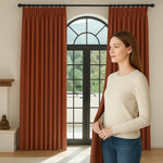 Femme en pull beige touchant un rideau marron plissé devant une grande fenêtre arquée avec vue sur la nature.