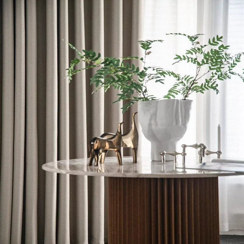 Décoration intérieure élégante avec une table ornée d'un vase blanc sculptural, contenant des plantes vertes, et des figurines dorées, devant de longues rideaux neutres. Ambiance contemporaine et raffinée, parfaite pour un salon moderne.