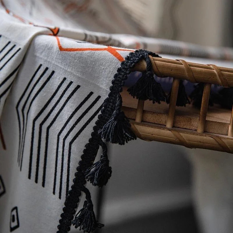 Plaid élégant avec motifs géométriques noirs et orange et franges décoratives noires, drapé sur un fauteuil en bois pour un intérieur moderne et chaleureux. Idéal pour ajouter une touche de style et de confort à votre salon ou chambre. Accessoire décoratif tendance pour une ambiance cosy.