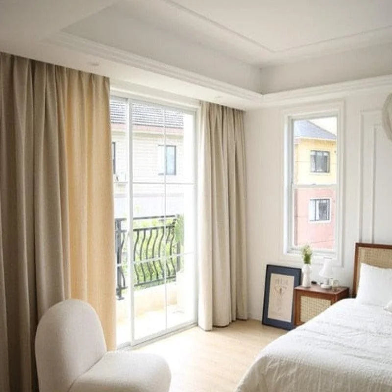 Chambre lumineuse et élégante avec de longs rideaux beiges couvrant une grande baie vitrée. Cette pièce confortable et moderne dispose d’un fauteuil design, d’un lit simple avec une élégante parure blanche et d’une table de chevet. Idéale pour créer une ambiance apaisante et chaleureuse dans votre intérieur.