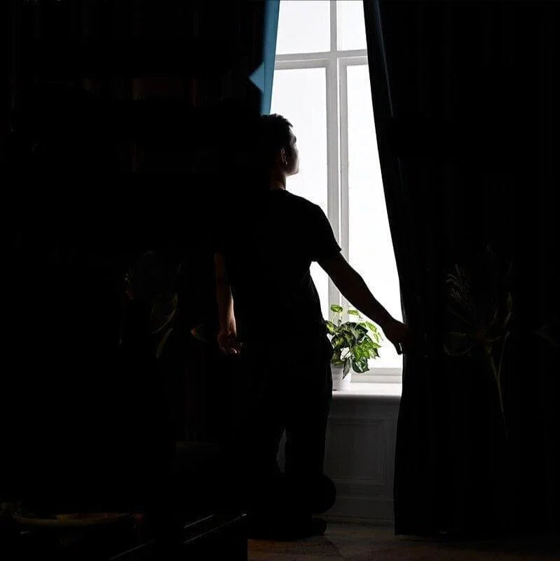 Homme devant une fenêtre ouverte, ajustant un rideau pour laisser entrer la lumière naturelle dans la pièce, avec une plante d'intérieur sur le rebord de la fenêtre, créant une ambiance de tranquillité et de bien-être à domicile.