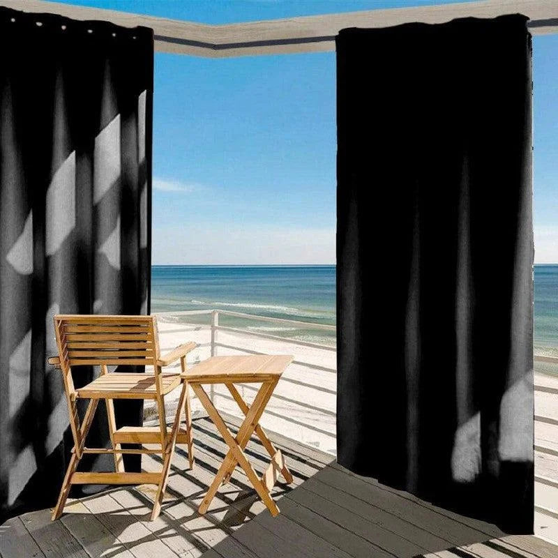 Rideaux occultants noirs suspendus sur une terrasse ensoleillée avec vue sur la mer, créant un espace intime et ombragé. Ensemble table et chaise en bois pour profiter du paysage côtier. Accessoire idéal pour décorer les balcons ou terrasses tout en préservant l'intimité.