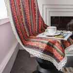 Couverture décorative colorée à motifs géométriques sur une chaise, agrémentée d'une bordure en dentelle, idéalement disposée près d'une cheminée pour créer une atmosphère chaleureuse. Accessoirisée avec une tasse de thé et un magazine, elle ajoute une touche bohème et élégante à votre intérieur.