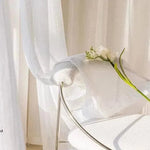 Chaise design moderne en métal, accentuée par un rideau léger et délicat. Un bouquet de fleurs blanches apporte une touche naturelle à cette ambiance lumineuse et épurée, parfaite pour décorer un intérieur contemporain.