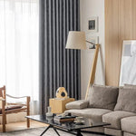 Salon moderne avec canapé gris et rideaux sombres, aménagé avec une lampe sur pied élégante et une table basse noire, accompagné d'une chaise contemporaine. Décoration intérieure chic avec accents de bois clair et lumière naturelle abondante. Idéal pour un espace de vie confortable et stylé.
