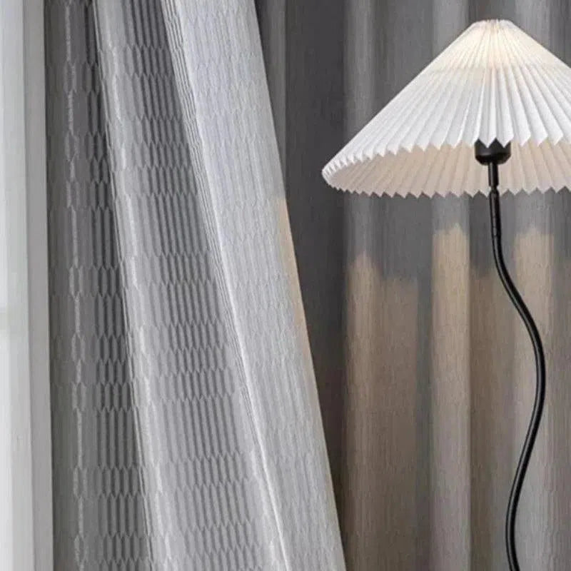 Lampe design avec abat-jour plissé blanc, idéale pour un éclairage élégant et moderne, située à côté d'un rideau texturé gris. Parfait pour une décoration intérieure raffinée.