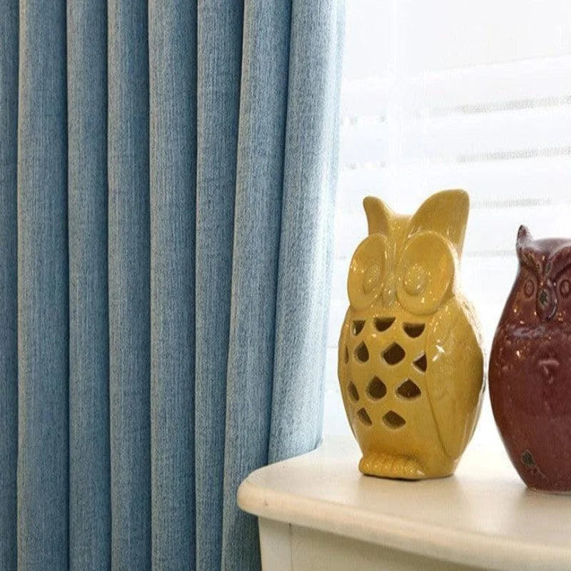 Rideau élégant bleu pour salon avec figurines décoratives en céramique en forme de hibou sur une table. Idéal pour ajouter une touche de style et d'originalité à votre décoration intérieure tout en modifiant l'ambiance lumineuse de la pièce. Accessoire incontournable pour une maison moderne et accueillante.