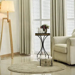 Salon élégant avec un fauteuil confortable et un tapis rond beige, illuminé par une lampe sur trépied. Table d'appoint noire avec décorations florales, devant une fenêtre avec des rideaux et des stores. Ambiance lumineuse et moderne, idéale pour un intérieur contemporain.